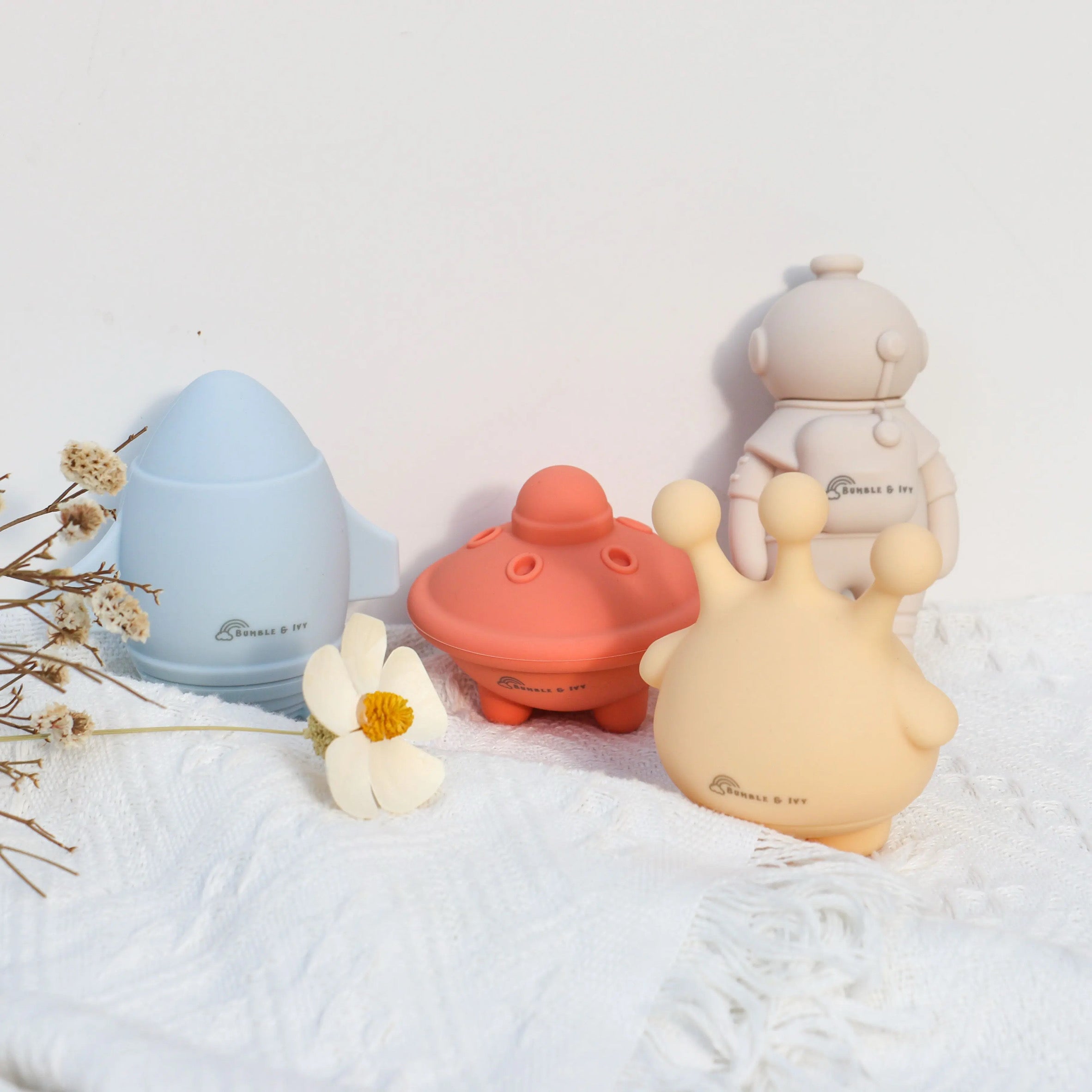 Bumble & Ivy mould-free silicone bath toys in a space theme, featuring a pastel blue rocket, peach UFO, yellow alien, and beige astronaut arranged elegantly on a white textured blanket with a flower.