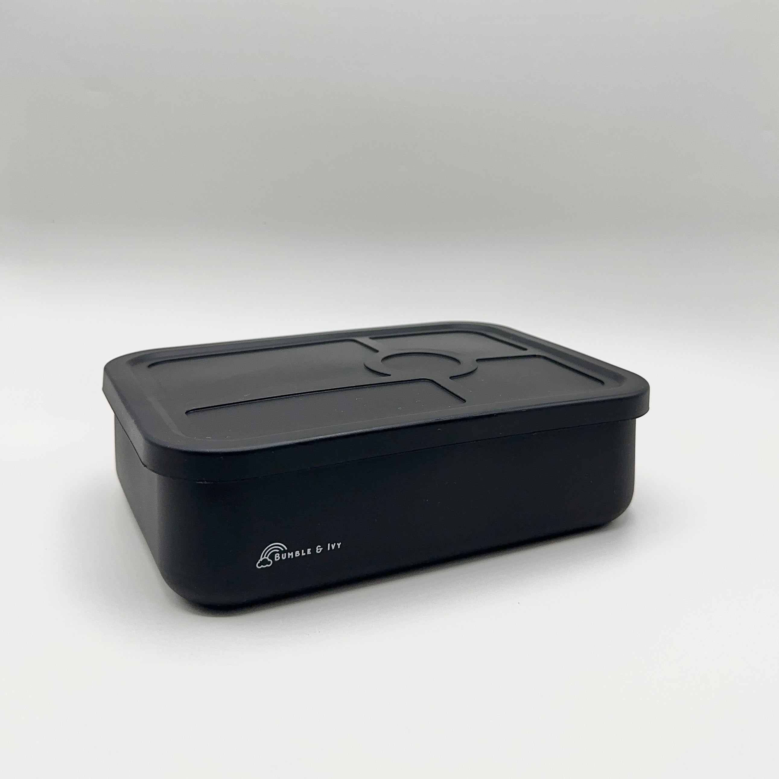 An angled view of the closed Bumble & Ivy midnight black silicone bento box on a white background, showing its durable, leakproof design and sectioned lid for kids' school lunches.