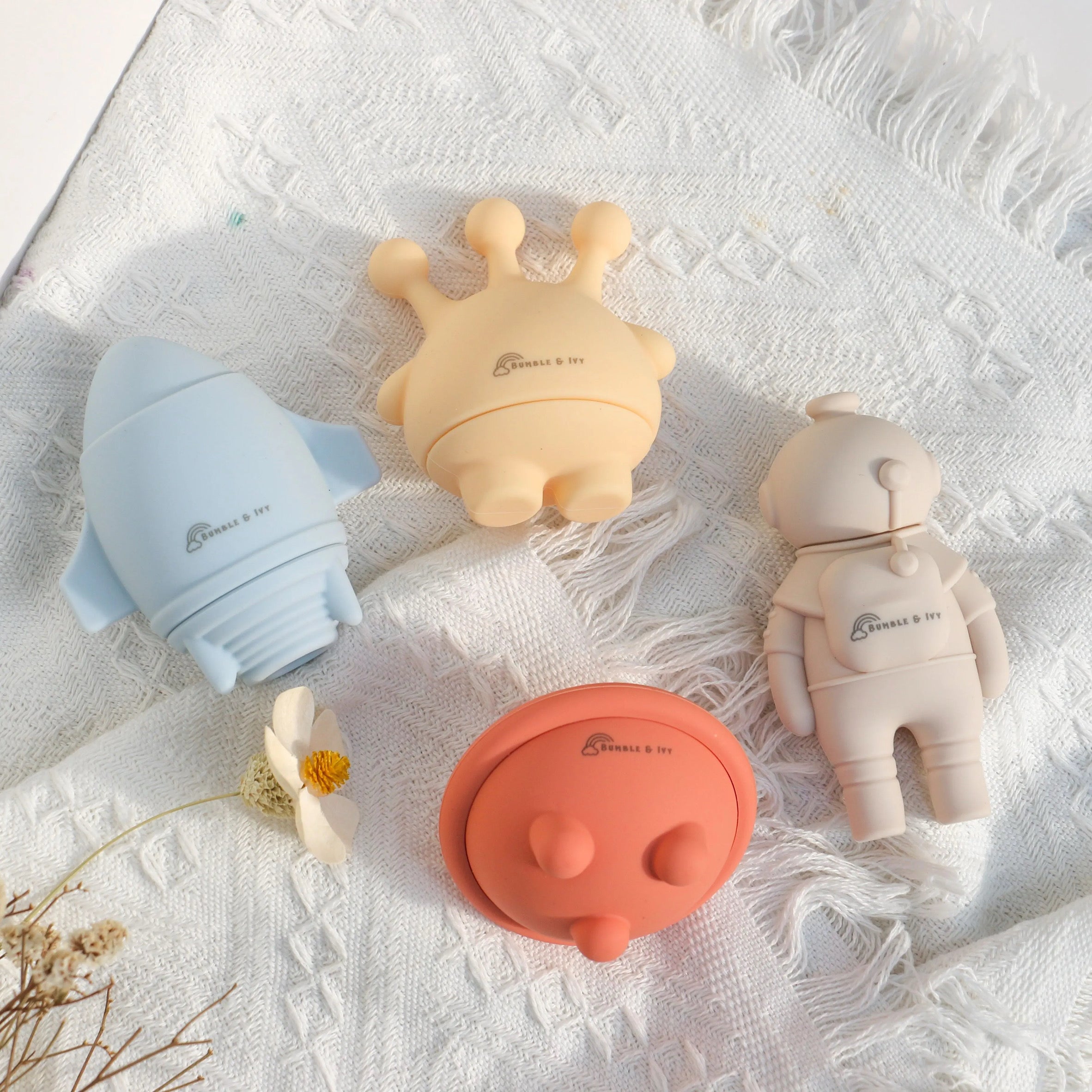 Top-down view of Bumble & Ivy easy-clean silicone bath toys, including a blue rocket ship, yellow alien, beige astronaut, and orange UFO neatly arranged on a white blanket.