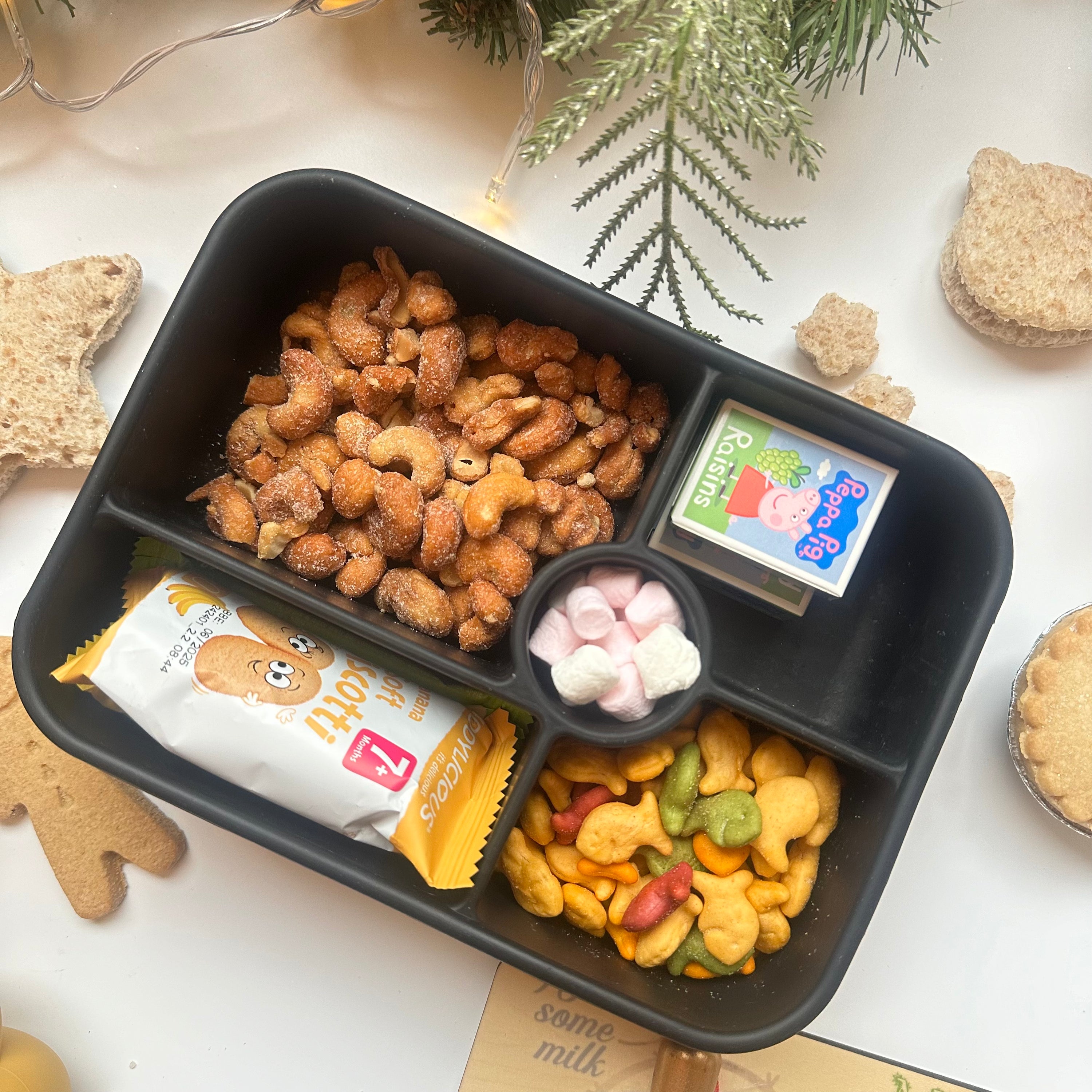 Overhead lifestyle shot of a Bumble & Ivy midnight black silicone bento box filled with Christmas snacks, including gingerbread biscuits, star-shaped sandwiches, raisins, and crackers, surrounded by festive holiday decorations.