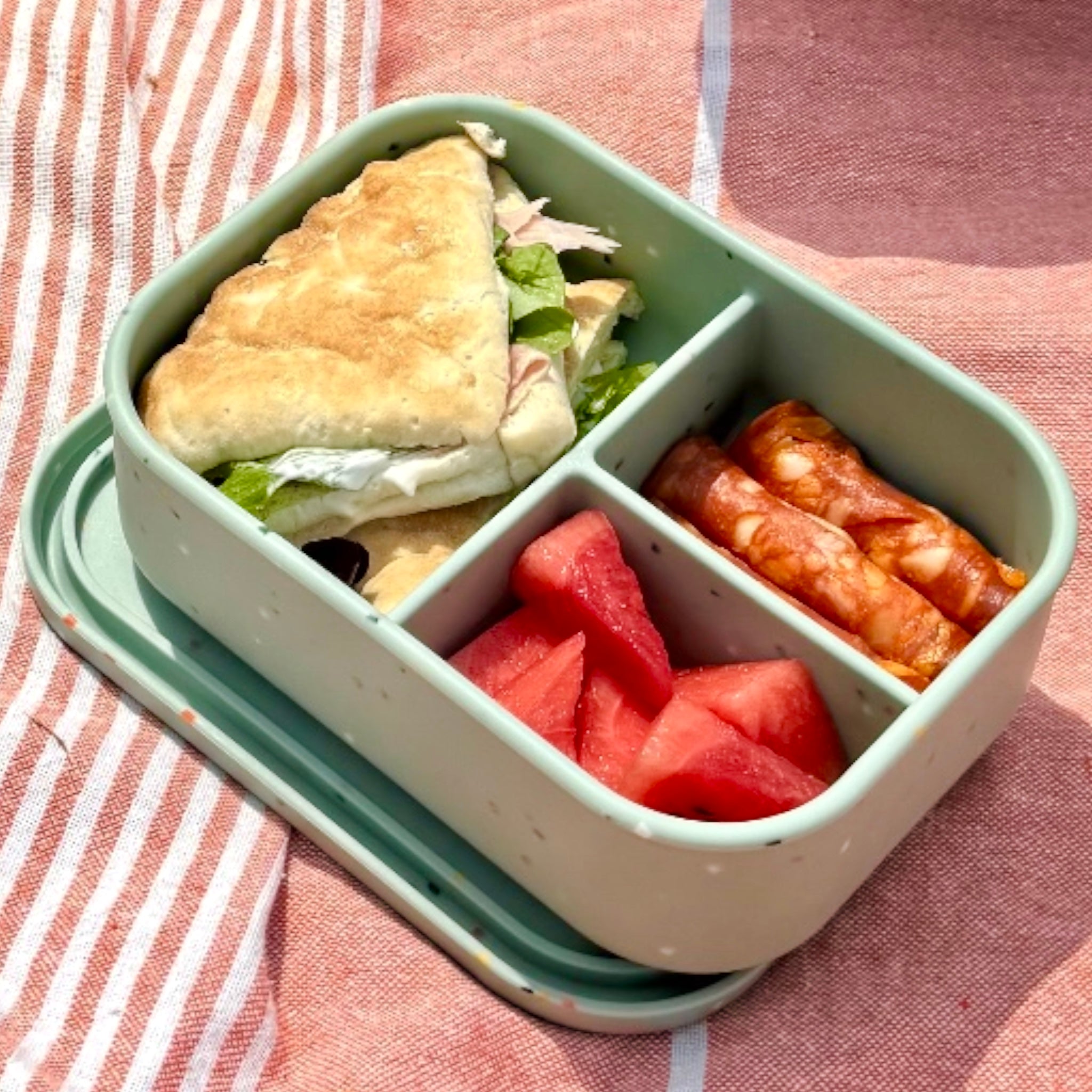 Bumble & Ivy Speckled Sage 3 compartment silicone bento lunch box filled with sandwich, watermelon and snacks on a striped picnic blanket. Leak-resistant lunch box for toddlers, school lunches, family days out and outdoor adventures